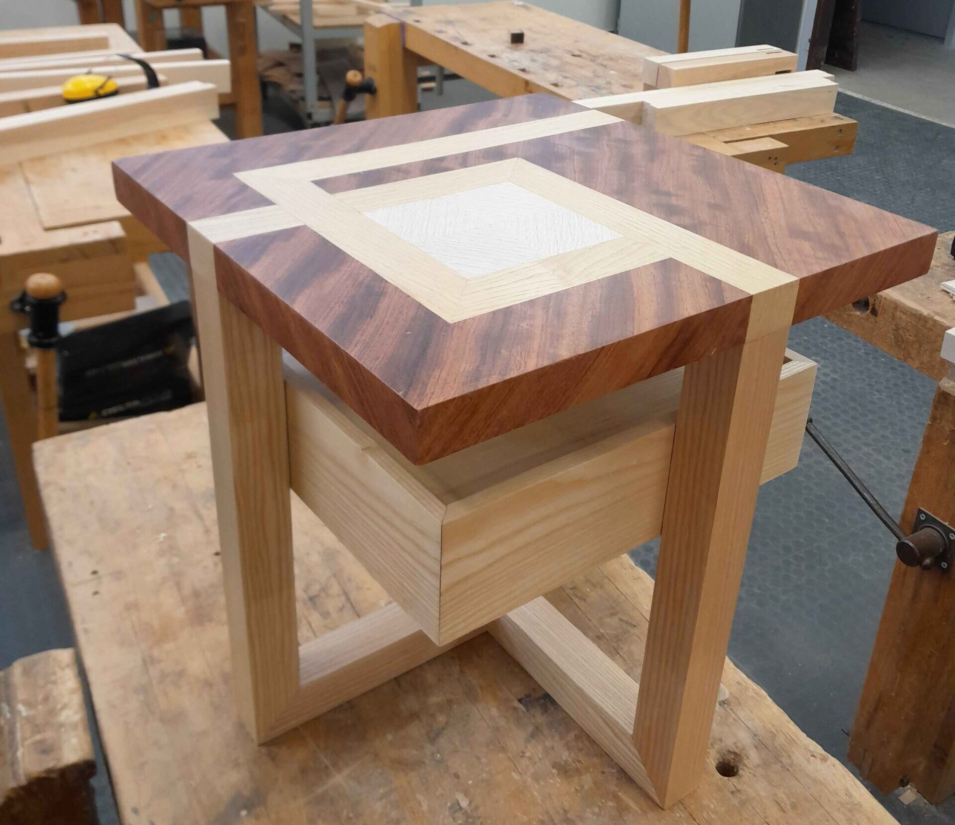 Photo d'une petite table au design cubique. Elle est formée d'un plateau cubique, d'un tiroir suspendu quelques centimètres sous le plateau et d'un piètement. Le plateau est marquété avec des bandes de la même couleur que les pieds, comme si les pieds se prolongeaient sur le plateau.