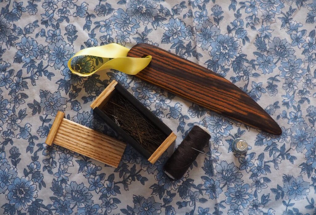 Photo d'un kit de couture vu du dessus, il y a une boite à aiguilles en ébène et en frêne et un plioir d'un bois d'un marron chaud aux veinage très marqué