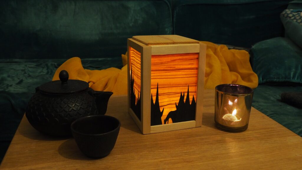 Une veilleuse en bois. C'est un cube et chaque face est faire d'un cadre qui tient une feuille de placage. Le placage très fin laisse passer la lumière depuis l'intérieur de la veilleuse ce qui révèle un veinage et des couleurs intenses. Sur chaque face une deuxième couche de placage empêche la lumière de passer, dessinant une forêt de sapins en négatif. Cette fois-ci, la veilleuse est dans la pénombre et on voit mieux la lumière passer à travers les panneaux.