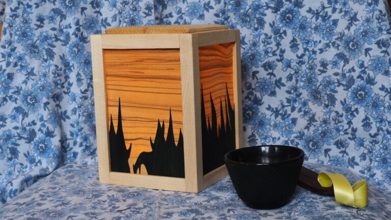 Une veilleuse en bois. C'est un cube et chaque face est faire d'un cadre qui tient une feuille de placage. Le placage très fin laisse passer la lumière depuis l'intérieur de la veilleuse ce qui révèle un veinage et des couleurs intenses. Sur chaque face une deuxième couche de placage empêche la lumière de passer, dessinant une forêt de sapins en négatif.