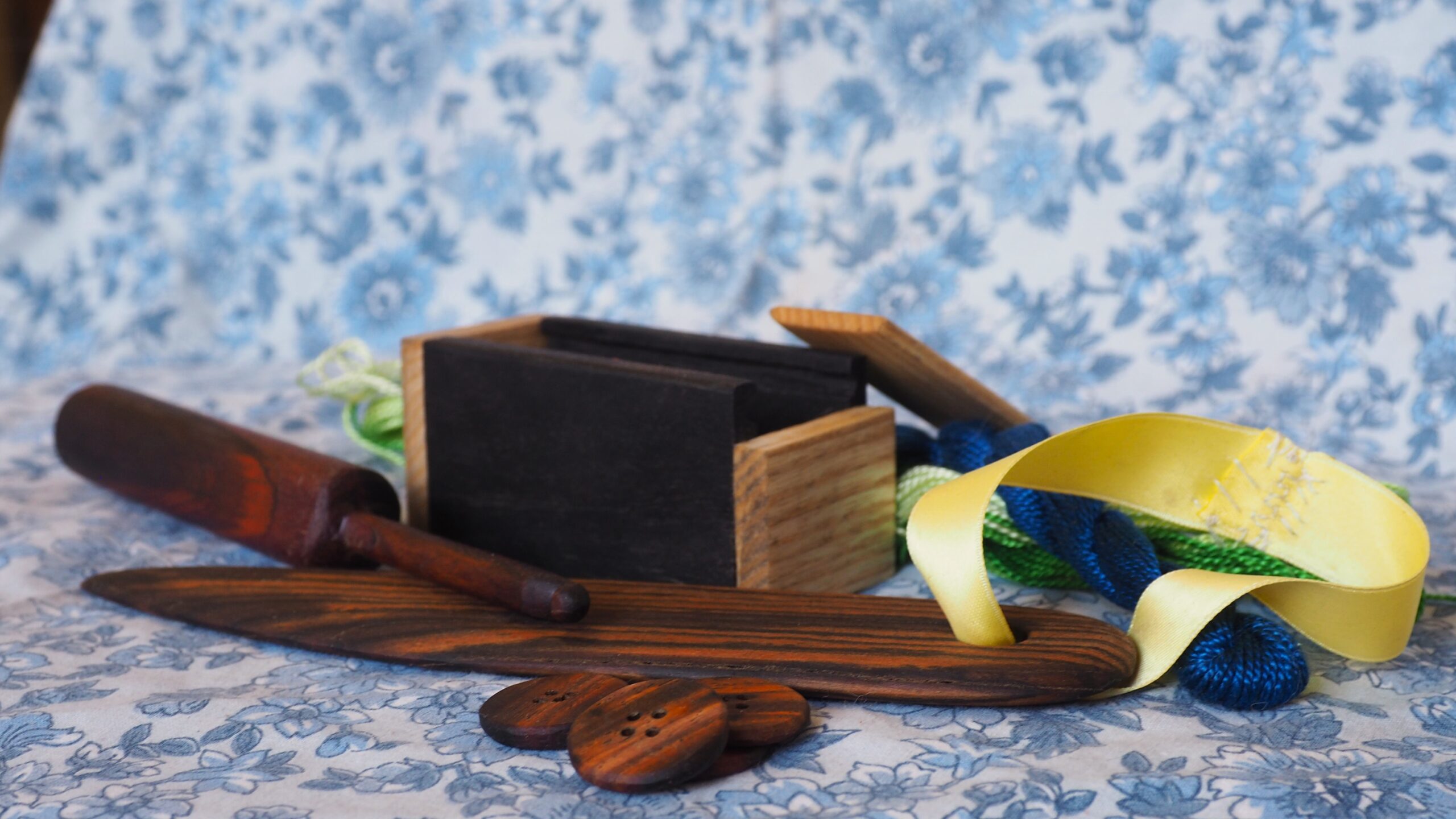 Différents objets en bois pour coudre posés sur un tissu à motif floral. Il y a un crochet, une boite à aiguilles, un long objet pour former les angles de coutures et des boutons en bois. Tous les objets sont faits avec des bois de couleurs différentes.